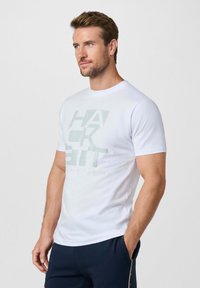 Hombre con barba y cabello corto, vestido con una camiseta blanca con estampado y pantalones oscuros, con una mano en el bolsillo, mirando hacia la derecha sobre un fondo liso.