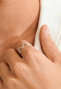Valoviti srebrni prsten nosi se na ruci, ima glatku teksturu i polirani završetak, s tankim, minimalističkim dizajnom i otvorenim krajevima.