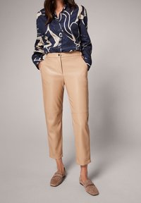 Donkerblauwe, gestippelde blouse met lange mouwen en abstracte ontwerpen. Beige, faux leren, taps toelopende broek met voorzakken en naden. Bruine instapschoenen.