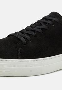 Czarny zamszowy sneaker z ciemnymi sznurówkami i białą gumową podeszwą. Subtelna faktura i detale szwów podkreślają minimalistyczny design.