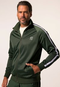 Chaqueta de chándal verde con cremallera, hecha de una tela suave, con acentos de rayas blancas y un logo en el lado izquierdo del pecho. Bolsillos a cada lado.