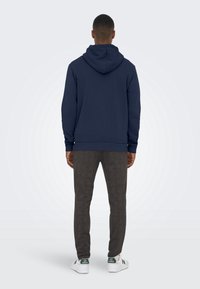 Felpa con cappuccio blu navy, dalla vestibilità aderente e con polsini a costine; abbinata a pantaloni scuri con fantasia e sneakers bianche con dettagli verdi.
