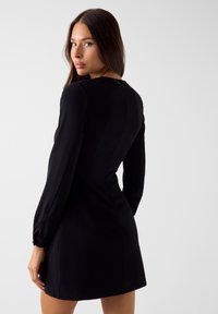 Schwarzes Minikleid mit langen Ärmeln, glattem Stoff, figurbetontem Design, Rückenreißverschluss und seitlichen Schlitzdetails.