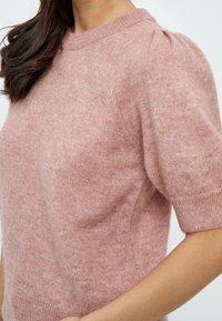 Žena v měkkém růžovém svetru s krátkým rukávem, vyklenutými rameny a žebrovaným výstřihem a manžetami, na jednobarevném světlém pozadí.