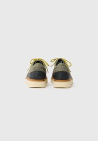 Pair of low-top lace-up shoes with green suede, black heel panels, yellow laces, and thick cream-colored soles, viewed from the back.