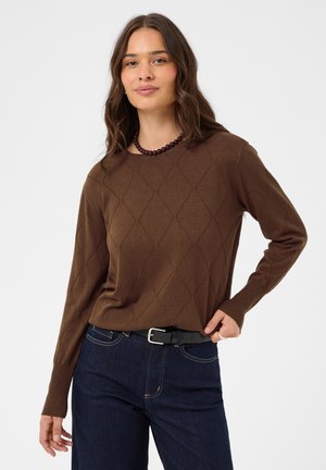 Femme aux longs cheveux bruns portant un pull marron à motif losange, un jean foncé, une ceinture noire et un collier de perles, debout devant un fond uni.