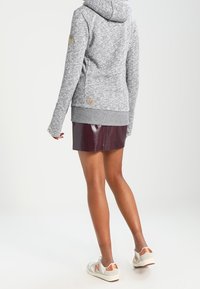 Grå huvtröja med texturerad yta och huva, kombinerad med en mörkt vinröd glansig kjol. Vita sneakers med bruna detaljer.