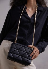 Sac à main noir matelassé avec un motif géométrique, bandoulière en chaîne dorée et poignée supérieure. Le matériau semble être en cuir texturé.