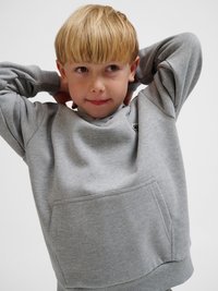 Enfant blond aux cheveux courts portant un sweat-shirt gris, mains derrière la tête, regardant légèrement sur le côté sur un fond uni.