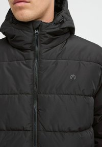 Melnās pūkainās jaka ar rāvējslēdzēja priekšpusi, regulējamu kapuci un quilted dizainu. Uz krūtīm ir diskrets logo. Gluda tekstūra.