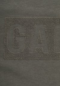 Textielstof in donkergrijs met gestructureerde, raised letters die "GAD" spellen. Gladde, gebreide oppervlakte met een subtiel weefpatroon.
