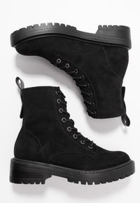 Botines de ante negro con suela de goma gruesa, diseño de cordones y tirador en la parte posterior. Superficie texturizada y detalles de costura.