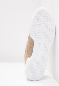 Semelle en caoutchouc blanche avec motif adhérent texturé sur une sneaker beige en mesh, positionnée verticalement contre un mur blanc d’angle.