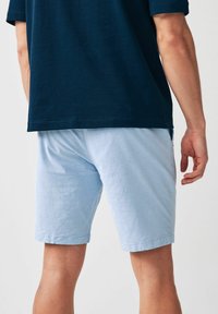 Helle blaue Baumwollshorts mit lässiger Passform, die über eine leichte Textur und einen Bund verfügen. Getragen mit einem dunkelblauen Hemd, von hinten betrachtet.