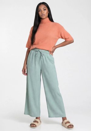 Kobieta ubrana w pomarańczowy sweter z krótkim rękawem i golfem, luźne miętowo-zielone spodnie w cienkie prążki oraz beżowe sandały na klamry, stoi z ręką na biodrze.