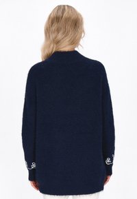Pulover tricotat de culoare bleumarin, cu gât înalt, croi lejer și mâneci lungi; prezintă accente brodate albe aproape de manșete. Material moale, texturat.