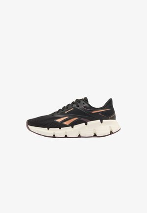 Schwarzer Reebok Laufschuh mit metallisch kupferfarbenen Akzenten und dicker weißer segmentierter Sohle, von links auf weißem Hintergrund gezeigt.