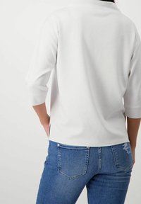 Pullover bianco con maniche a tre quarti, caratterizzato da spacchi laterali. Abbinato a jeans in denim blu, che mostrano un dettaglio sfilacciato sulla tasca posteriore.