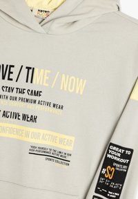 Grijze hoodie met gele accenten, voorzien van opvallende tekstontwerpen. Gemaakt van katoen, met een buidelzak, zachte textuur en een verstelbare capuchon.