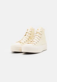 Korkeat sneakerit cremevärisellä kankaalla, joissa on valkoiset nauhat, kultaiset silmukkaketjut ja teksturoitu kumipohja, jossa on kuvioitu reuna.