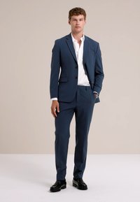 Jeune homme debout, portant un costume bleu marine avec une chemise blanche, des chaussures noires habillées et une montre, devant un fond beige uni.