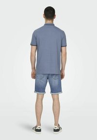 Camisa polo azul con cuello, combinada con pantalones cortos de mezclilla azul claro. Los pantalones cortos tienen dobladillos enrollados y bolsillos traseros. Zapatillas negras y blancas.