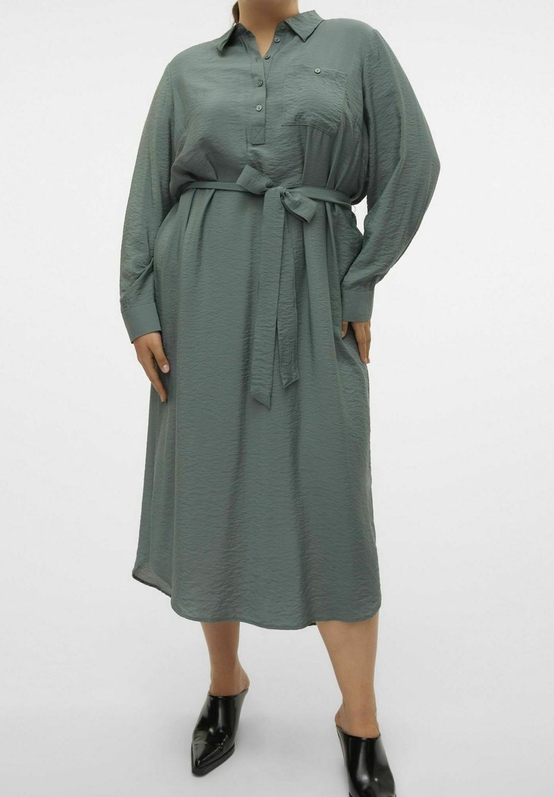 Personne portant une robe verte texturée à manches longues, boutonnée, avec une ceinture à la taille et des chaussures noires à bouts pointus sans lacets.