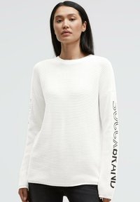 Weißer Strickpullover mit geripptem Muster, rundem Ausschnitt und langen Ärmeln mit schwarzem Text entlang des linken Arms. Hintergrund ist hellgrau.