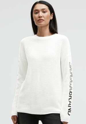 Weißer Strickpullover mit geripptem Muster, rundem Ausschnitt und langen Ärmeln mit schwarzem Text entlang des linken Arms. Hintergrund ist hellgrau.