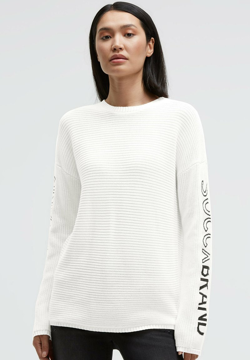 Weißer Strickpullover mit geripptem Muster, rundem Ausschnitt und langen Ärmeln mit schwarzem Text entlang des linken Arms. Hintergrund ist hellgrau.