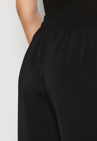 Svarta byxor med elastiskt midjeband som har en mjuk och slät kvalitet med subtila veck och en avslappnad passform, vilket framhäver en enkel och minimalistisk design.