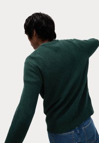 Dunkelgrüner strukturierten Pullover mit rundem Halsausschnitt und langen Ärmeln, getragen zu blauen Jeans. Das Material scheint gestrickt mit einem dezenten Muster zu sein.