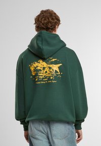 Grøn hoodie med et stort gult grafik, der viser en scene med dyr. Har en hætte med snoretræk og en løs pasform. Fremstillet af blødt stof.