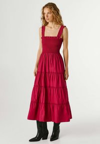 Moteris dėvi raudoną, berankovę, sluoksniuotą suknelę su plačiomis petnešėlėmis ir juodus puskojinio ilgio aukštakulnius batus, stovinti priešais paprastą fono sieną.