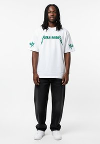 T-shirt blanc à manches courtes avec un graphisme vert "PARIS" et un design d'étoiles sur les manches, associé à un jean en denim foncé et des baskets blanches.