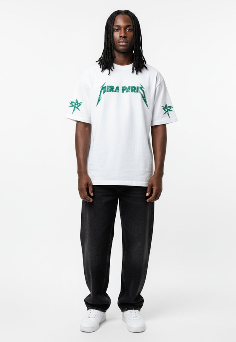 T-shirt blanc à manches courtes avec un graphisme vert "PARIS" et un design d'étoiles sur les manches, associé à un jean en denim foncé et des baskets blanches.