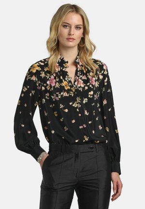 Femeie blondă stă cu mâna în buzunar, purtând o bluză neagră cu flori și mâneci lungi și pantaloni negri eleganți, pe un fundal simplu.