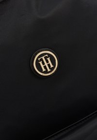 Badge rond noir et or avec le logo Tommy Hilfiger sur un tissu noir lisse, avec une texture polie et des coutures discrètes visibles au bord inférieur.