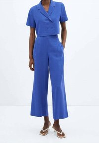 Donna che indossa un blazer cropped doppiopetto a maniche corte di colore blu cobalto e pantaloni a gamba larga abbinati, completati con sandali con tacco bianchi.