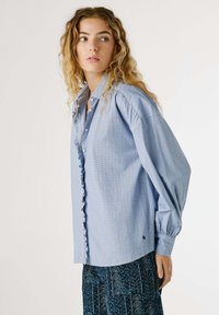 Himmelblaues Hemd mit Knopfleiste, strukturiertem Muster, Rüschenapplikation entlang der Knopfleiste und weit geschnittenen Ballonärmeln.