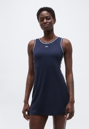 Mujer con vestido deportivo sin mangas azul marino con ribetes blancos y logo en el pecho, de pie frente a un fondo claro y liso.