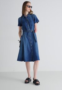 Denim shirt dress with short sleeves, buttoned front, belted waist, and flared skirt. Dark blue color, smooth texture, paired with platform sandals.