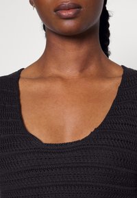 Top en tricot noir avec un col arrondi, présentant un motif texturé et une coupe ajustée qui met en valeur les clavicules.