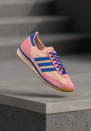 Sneaker rosa e beige con strisce blu, materiali in suede e lisci, punta rotonda e area delle stringhe testurizzata, su sfondo grigio.