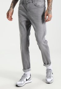 Jeans in denim grigio chiaro con una vestibilità slim, con orli arrotolati e tasche frontali, abbinati a sneakers Nike bianche e grigie con dettagli neri.