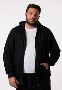 Felpa nera con zip, dotata di cappuccio regolabile, caratterizzata da un tessuto testurizzato e un piccolo logo sul petto. Indossata sopra una t-shirt bianca.