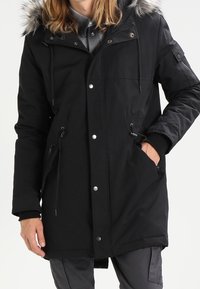 Svart parkas med huva av päls, dragkedja och tryckknappsstängning, två sidofickor, justerbara dragsnören och strukturerad tyg.