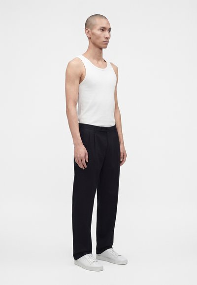 Homme debout, face à droite, portant un débardeur blanc sans manches, un pantalon noir plissé et des baskets blanches sur un fond clair uni.