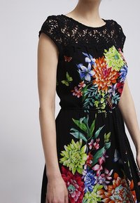 Blumenkleid aus schwarzem, fließendem Stoff. Es zeigt bunte Blumenmuster, einen gehäkelten Spitzen-Ausschnitt und eine taillierte Detailgestaltung.