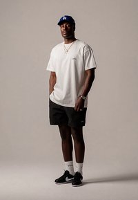 Man som bär vit t-shirt, svarta shorts, svarta sneakers, vita strumpor, blå basebollkeps och guldkedjor, står mot en enfärgad bakgrund.